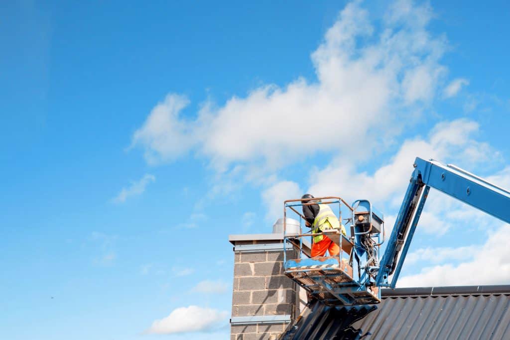A worker stands on a blue lift, expertly performing chimney repair against a clear blue sky with scattered clouds. Wearing a neon safety vest and orange pants, they focus intently on the task. The lift is positioned beside the building's sloped roof.