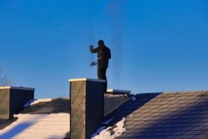 A person stands on a snowy rooftop, meticulously performing chimney cleaning. The clear blue sky contrasts with the dark roof and chimney caps, while shadows cast by the setting or rising sun highlight this serene scene.