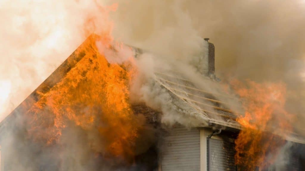 A house engulfed in flames and thick smoke, with the roof partially collapsed and charred. Intense orange flames and dark gray smoke billow from the structure, suggesting a severe fire possibly ignited by neglected chimney repair needs.