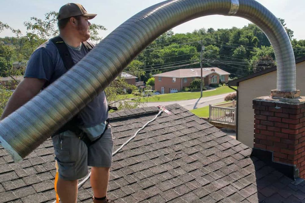 A person on a roof, wearing work gear, is handling a large, flexible metal duct connected to a brick chimney. They seem to be assessing the area for possible chimney repair. The background shows a residential neighborhood with trees and houses.